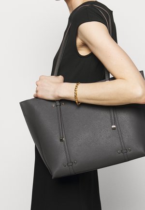 Donna che indossa un vestito nero senza maniche, con una grande borsa a tracolla grigia scura e texture, bracciale in oro al polso.