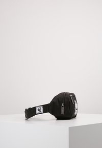 Banane en tissu noir de forme ronde, dotée d'une poche avant zippée, d'une sangle ajustable et d'accents de logo blanc.