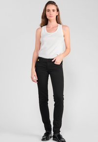 Jean skinny noirs avec des accents de boutons dorés, associés à un haut sans manches blanc, présentés sur un mannequin contre un fond uni.