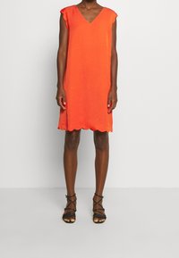 Korte oranje jurk met een V-hals en een gegolfde zoom, voorzien van korte gerafelde mouwen en een losse pasvorm. Gestyled met zwarte sandalen met bandjes.