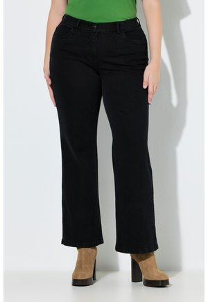 Person trägt schwarze Jeans mit geradem Bein, ein grünes Oberteil und beigefarbene Wildleder-Stiefeletten mit hohem Absatz, steht vor einem weißen Hintergrund.