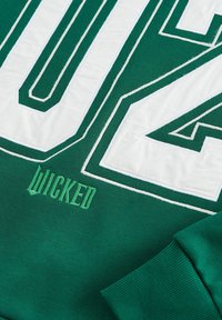 Zelena dukserica s velikim bijelim brojevima; "WICKED" izvezena u zelenoj boji. Mekane teksture s rebrastim manžetama i ovratnikom.