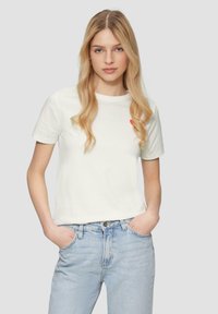 Witte katoenen t-shirt met korte mouwen, ronde hals en een klein rood hartgrafiekje op de linker borst. Gecombineerd met lichtblauwe denim jeans.