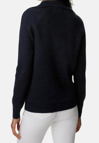 Marineblauwe gebreide trui met een ribbelstructuur, voorzien van een klassieke kraag en lange mouwen, gecombineerd met witte jeans.