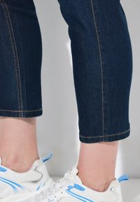 Tummansinisiä farkkuja, joissa on lyhennetty helma, näkyvillä ompelusaumoin, yhdistetty valkoisiin lenkkareihin, joissa on sinisiä ja hopeisia yksityiskohtia.