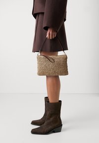 Borsa a tracolla intrecciata in paglia di forma rettangolare, colore beige, con manico in pelle marrone, texture dettagliata, tenuta in mano accanto a stivali alla caviglia in suede marrone.