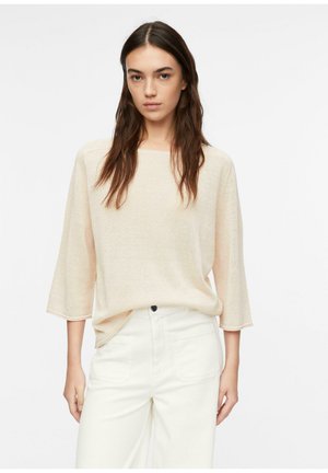 Frau mit langen braunen Haaren, die einen lockeren beigen Pullover mit Dreiviertelärmeln und eine hoch taillierte weiße Hose vor weißem Hintergrund trägt.