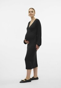 Abito premaman nero a coste con scollatura profonda a V e maniche lunghe, caratterizzato da una silhouette aderente che arriva a metà polpaccio. Scarpe nere basse.
