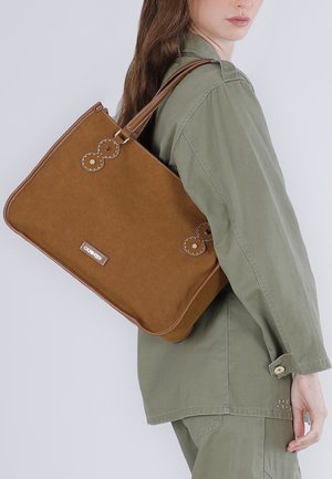 Donna che indossa una giacca e pantaloni verde oliva, con una grande borsa a tracolla marrone con dettagli cuciti sulla spalla.