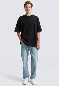 Jauns vīrietis stāv, ģērbies brīvā melnā T-kreklā, gaiši zilās džinsās un baltās kedās, ar vienu roku kabatā.