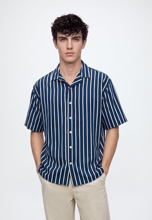 Jeune homme aux cheveux bouclés et foncés porte une chemise boutonnée à manches courtes à rayures verticales bleu et blanc et un pantalon beige, les mains dans les poches.