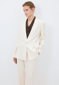 Ljus krämfärgad kostym med randigt mönster, bestående av en strukturerad kavaj och vida byxor. Kavajen har två framfickor och en enkel knappstängning.