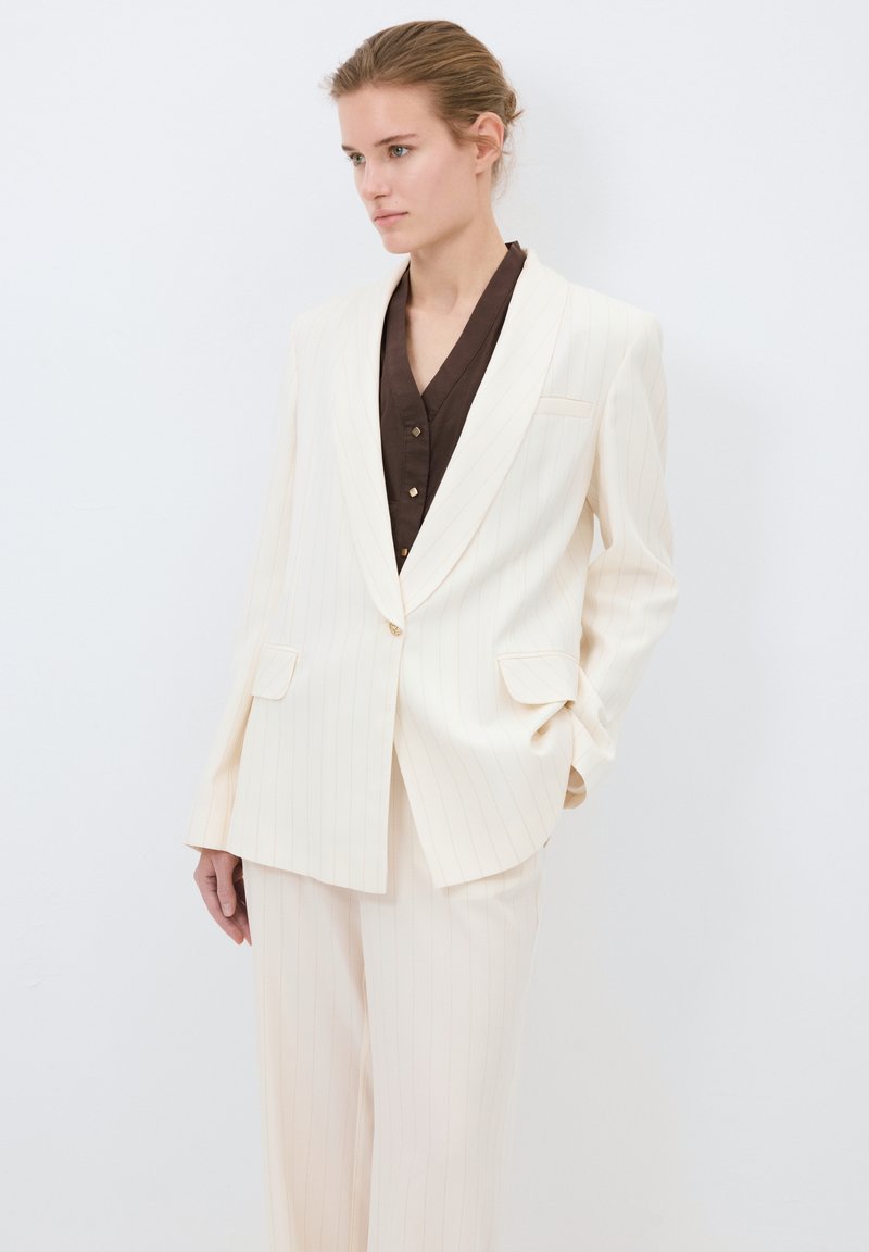 Ljus krämfärgad kostym med randigt mönster, bestående av en strukturerad kavaj och vida byxor. Kavajen har två framfickor och en enkel knappstängning.