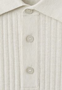 Polo en maille beige clair avec col et trois boutons sur le devant, présentant une texture côtelée verticale.