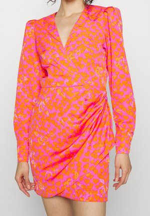 Kvinde iført en rosa slå-om-kjole med lange ærmer og et orange hjerte mønster, med rynkede detaljer på nederdelen, stående mod en ensfarvet baggrund.