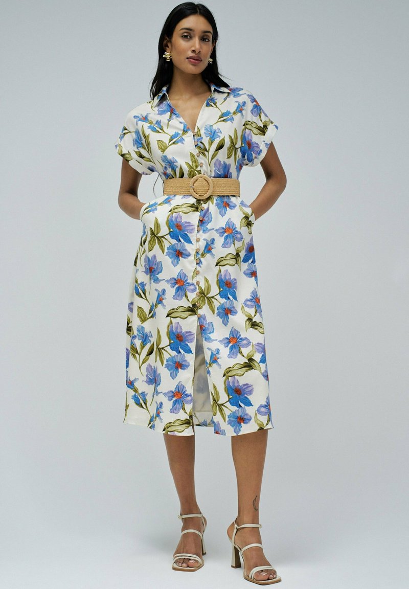 Vestido midi floral en blanco, con flores azules y naranjas, mangas cortas, frente de botones y un cinturón trenzado beige en la cintura.