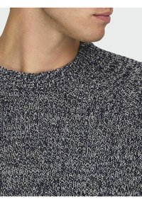Navyblauwe gebreide trui met witte stippen, met een ribgebreide ronde hals en een textuurpatroon, waarop een close-up van de stof te zien is.