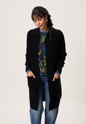 Vrouw die een lange zwarte cardigan draagt over een gebloesd shirt en blauwe spijkerbroek, met haar handen in de zakken van de cardigan en een gevlochten kapsel.