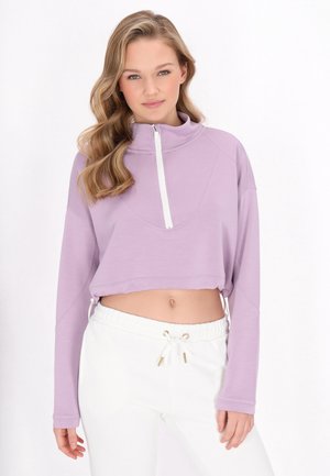 Lavendel cropped sweatshirt med høj krave og kvart længde lynlås, parret med hvide bukser med snor i livet og guld accenter.