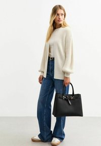 Cardigan lavorato a maglia color crema, top grigio chiaro, jeans blu a vita alta, borsa nera con accessori in metallo, sneakers bianche, posa in piedi.