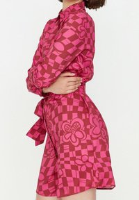 Dlouhá halenka v růžové barvě s kostkovaným vzorem a květinovými motivy. Obsahuje límeček a detail s uvázaným pasem. Vyrobeno z lehkého materiálu.