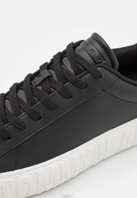 Černé kožené tenisky s černými tkaničkami a strukturovanou bílou gumovou podrážkou. Obsahují logovou nášivku na jazyku a hladký kožený svršek.