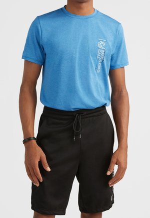 Homme portant un t-shirt graphique bleu avec le texte "Santa Cruz Cali" et un short noir à cordon, debout devant un fond uni.