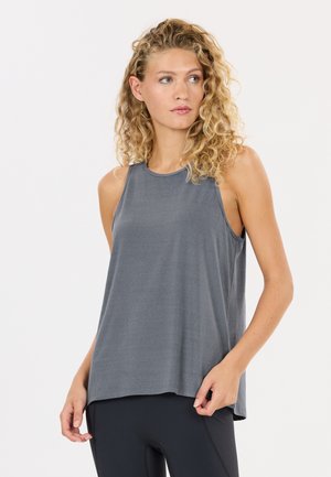 Vrouw met krullend blond haar, gekleed in een losse grijze mouwloze top en zwarte legging, kijkt iets naar opzij tegen een witte achtergrond.