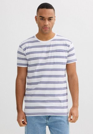 Hombre con camiseta blanca de manga corta con rayas horizontales azul marino y jeans azul claro, de pie contra un fondo blanco liso.