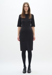 Černé přiléhavé šaty s krátkými rukávy, délka ke kolenům, hladká textura, minimalistický design, doplněné černými punčochami a šněrovacími koženými botami.