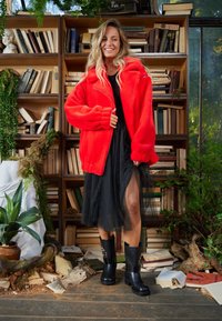 Piros, bársonyos cipzáras dzseki bordázott mandzsettákkal, fekete tüll szoknya fölött viselve. A modell fekete, vastag sarkú csizmát visel. A háttérben könyvespolcok láthatók.