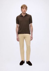 Jeune homme debout portant un polo marron en maille à manches courtes, un pantalon beige et des mocassins marron foncé sur un fond blanc uni.