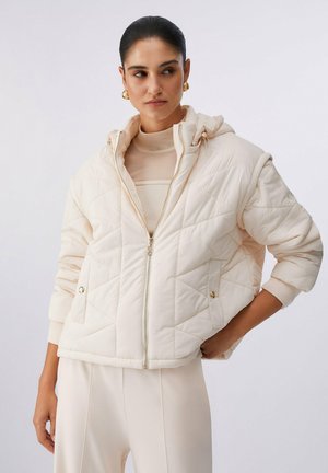 Frau trägt eine cremefarbene, gesteppte Jacke mit Kapuze, hohem Kragen und goldenen Knopfdetails, kombiniert mit passenden hellen Hosen und goldenen Ohrringen.