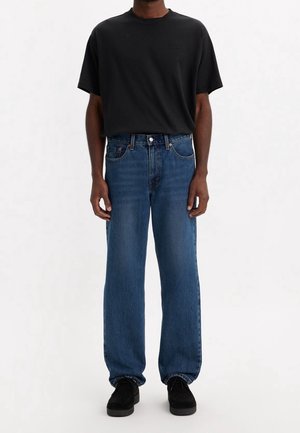 Personne portant un t-shirt noir uni à manches courtes rentré dans un jean bleu droit avec des chaussures noires, debout devant un fond blanc.