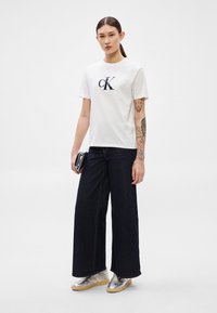 Valge puuvillane t-särk tumelilla logoga, kombineeritud laia säärega tumedate teksade ja metallhõbedaste tennistega. Modell hoiab musta kotti.