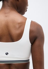 Ανώτερο μέρος της πλάτης και ο ώμος μιας γυναίκας που φοράει λευκό αθλητικό σουτιέν με μαύρη και λευκή ριγέ ταινία και μικρό μαύρο λογότυπο.