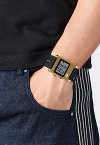 Skaitmeninis laikrodis su auksine spalva dengtu korpusu, juodu silikono dirželiu, kvadratine forma ir aiškiu LCD ekranu, rodantį laiką. Metalinės detalės palei kraštus.