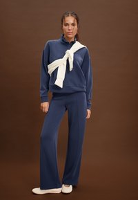 Marineblauer Pullover mit hohem Kragen, kombiniert mit passenden, weit geschnittenen Hosen. Cremefarbener Schal um den Hals gebunden; glatter Stoff und lockere Passform.