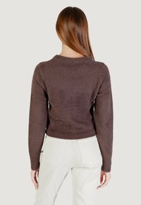 Donna con capelli castani lisci, che indossa un maglione lungo a maniche lunghe di colore marrone scuro e pantaloni bianchi a vita alta, in piedi con la schiena rivolta verso la telecamera.