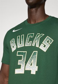 Žalias medvilninis marškinėliai su užrašu "BUCKS" ir "34" balta spalva su blizgesio efektu. Nike logotipas yra kairiajame petyje.