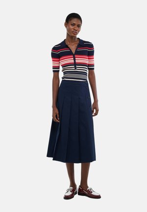 Femme portant une jupe plissée bleu marine avec des boutons sur le côté, un polo à manches courtes rayé bleu marine, rouge, blanc, et des mocassins bordeaux.