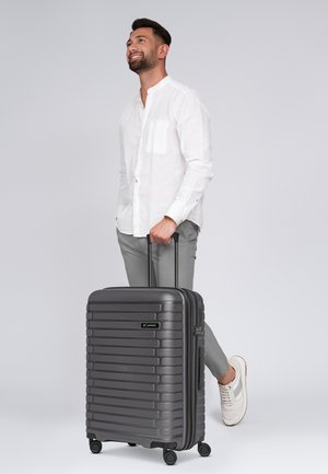 Valise rigide gris foncé avec des stries horizontales, poignée rétractable, quatre roues pour une maniabilité optimale et fermeture à glissière.