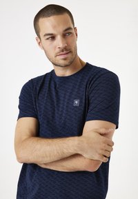 Camiseta de manga corta azul marino con un sutil patrón de olas, que presenta un pequeño parche de logo en el pecho y un cuello redondo.