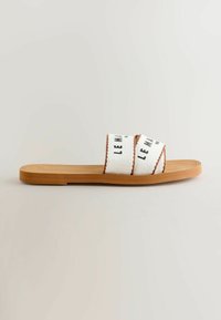 Sandale plate couleur beige avec deux lanières en tissu blanc croisées sur le dessus, ornées de texte noir et de détails de couture marron.