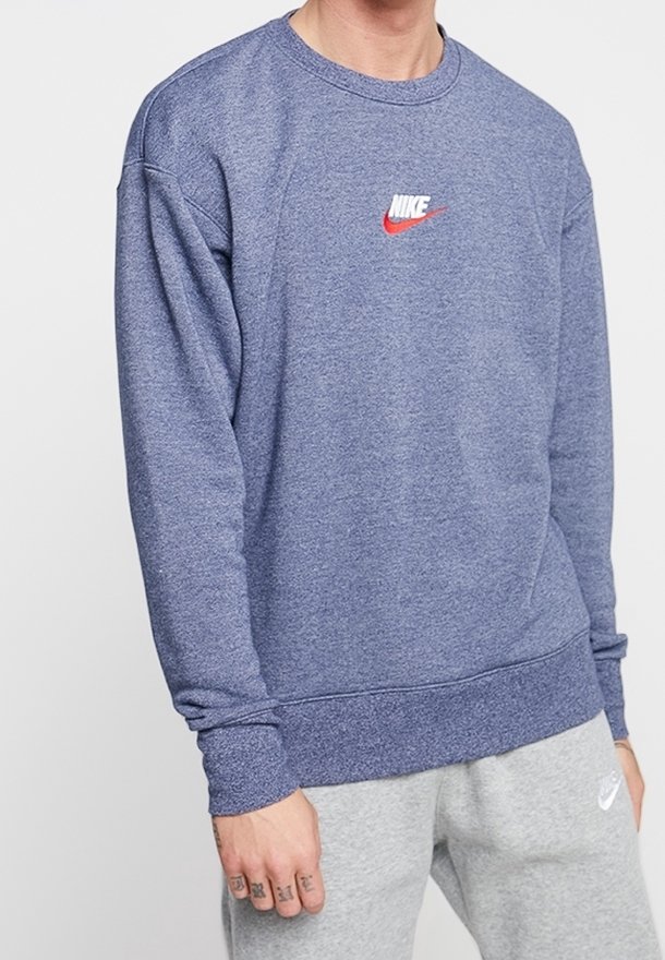 Personne portant un sweat-shirt Nike bleu avec un logo Swoosh rouge et un pantalon de jogging Nike gris clair, debout devant un fond uni.