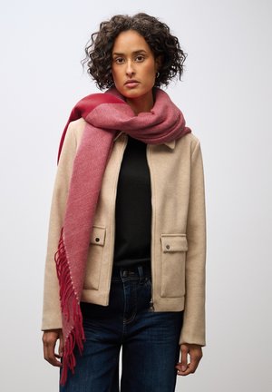 Veste en mélange de laine beige avec fermeture éclair et deux poches avant, associée à une écharpe douce rayée rouge et rose avec franges. Jean bleu.