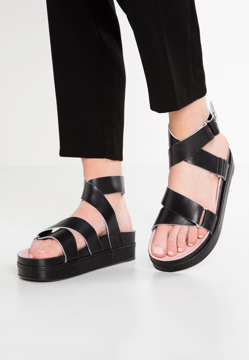 Černé kožené platformové sandály s širokými pásky, stříbrnými akcenty a zaobleným designem špičky. Obsahují nastavitelné spony pro bezpečné upevnění.