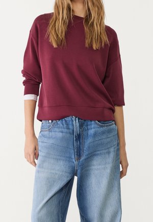 Vrouw draagt een bordeauxrode top met lange mouwen met opgerolde mouwen over een wit overhemd, gecombineerd met losse lichtblauwe spijkerbroek.