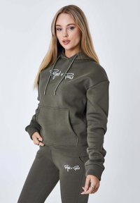 Felpa verde oliva realizzata in tessuto morbido, con tasca frontale, cappuccio con lacci e testo del logo bianco. Abbinata a pantaloni jogger coordinati.