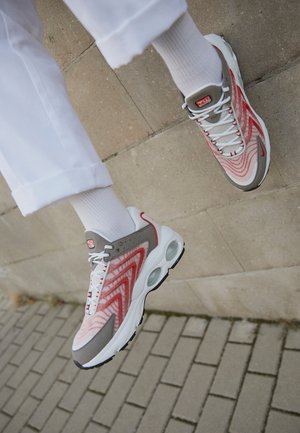 Personne portant un pantalon blanc et des chaussettes blanches avec des chaussures de sport rouges et grises flottant au-dessus d’un sol pavé.